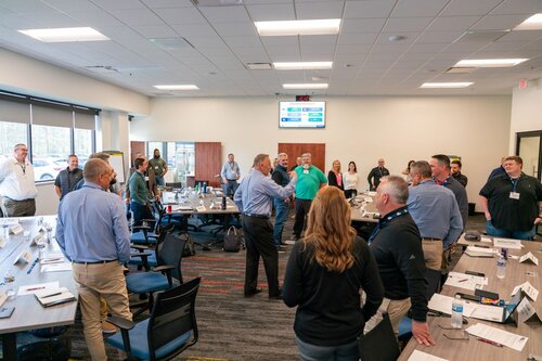 The image depicts a professional meeting or workshop setting in a modern conference room. Approximately 20 individuals are present, standing in a semi-circle around a central figure who appears to be leading a discussion or presentation. The participants are dressed in business casual attire, suggesting a formal yet relaxed atmosphere.

The room is well-lit with natural light streaming in from large windows on the left side, complemented by overhead fluorescent lighting. The walls are neutral, enhancing the professional ambiance. The floor is carpeted with a pattern that includes shades of gray and red, adding a subtle touch of color to the setting.

Tables are arranged in a U-shape, covered with papers, notebooks, and water bottles, indicating an interactive session where note-taking and hydration are encouraged. Laptops and other electronic devices are visible, suggesting the use of digital tools for the meeting.

On the wall at the front of the room, a digital screen displays information, possibly an agenda or presentation slides, though the specific content is not discernible. This setup implies a structured meeting with a clear focus.

The overall atmosphere is collaborative and engaged, with participants actively listening and interacting with the speaker. The setting and activities suggest a corporate training session, strategic planning meeting, or team-building workshop. The presence of diverse individuals indicates inclusivity and a focus on teamwork.

This image captures the essence of a dynamic and organized professional environment, emphasizing communication, learning, and collaboration.