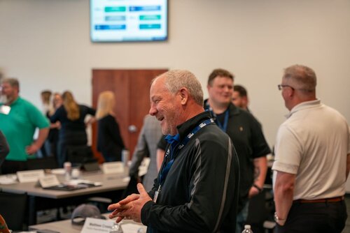 The image depicts a lively and informal meeting or conference setting. Several individuals are engaged in conversation, suggesting a networking or collaborative atmosphere. The focus is on a man in the foreground, who is smiling and appears to be speaking with others, indicating a positive and engaging interaction. He is wearing a black jacket and a lanyard, typical of conference or seminar attendees.

In the background, more people are gathered in small groups, some standing and others seated at tables. The tables are equipped with nameplates, papers, and water bottles, further emphasizing the professional and organized nature of the event. A large screen is visible on the wall, displaying a presentation slide with graphs or charts, suggesting a business or educational context.

The room is well-lit with neutral tones, creating a professional yet welcoming environment. The overall atmosphere is one of camaraderie and professional engagement, typical of workshops, seminars, or networking events.