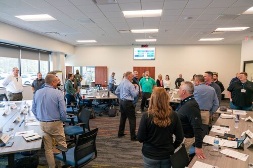 The image depicts a professional meeting or workshop setting in a modern conference room. The room is spacious and well-lit, with fluorescent ceiling lights and large windows on one side, allowing natural light to filter in. The atmosphere is collaborative and interactive, as evidenced by the group of approximately 20 individuals engaged in conversation and discussion.

The participants, both men and women, are casually dressed in business attire, such as collared shirts and slacks. They are standing around tables arranged in a U-shape, suggesting a participatory or workshop-style meeting. Each table is equipped with nameplates, water bottles, and papers, indicating an organized and formal setup.

Prominent objects in the room include several chairs, laptops, and presentation materials. A large screen at the front of the room displays a digital presentation or agenda, though the specific content is not clearly visible. The walls are neutral in color, contributing to a professional environment.

The carpet has a modern design with a mix of dark and light tones, adding a touch of color to the otherwise neutral setting. The overall mood is one of engagement and collaboration, typical of a business or educational seminar.

This setting is indicative of a corporate training session, a strategic planning meeting, or a workshop aimed at fostering teamwork and communication. The presence of diverse participants suggests inclusivity and a focus on group dynamics.