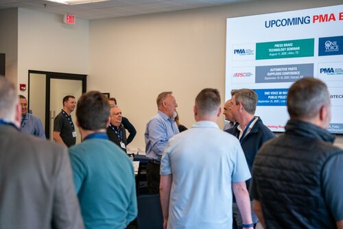 The image depicts a professional networking or conference setting, likely related to the manufacturing or technology industry. Several individuals, predominantly men, are engaged in conversation, suggesting a social or networking event. They are dressed in business casual attire, with some wearing name badges, indicating a formal gathering.

In the background, a large digital screen displays information about upcoming events. The screen features logos for organizations such as PMA (Precision Metalforming Association) and NTMA (National Tooling and Machining Association), along with event titles and dates. Events listed include a "Press Brake Technology Seminar" and an "Automotive Parts Suppliers Conference," suggesting a focus on industry-specific topics.

The room is well-lit with neutral colors, contributing to a professional and focused atmosphere. The presence of a table with papers and other materials suggests a seminar or workshop component to the event.

Overall, the image conveys a theme of professional development and industry collaboration, with an emphasis on upcoming educational opportunities in the manufacturing sector.