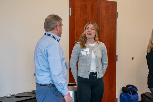 The image depicts two individuals engaged in conversation in what appears to be a professional or conference setting. The man on the left is wearing a light blue shirt and dark pants, with a lanyard around his neck, suggesting he might be attending an event or conference. The woman on the right is wearing a gray cardigan over a white top and dark pants. She has a name tag that reads "Heather," indicating her participation in the event.

The setting is indoors, with neutral-colored walls and a wooden door in the background, suggesting a conference room or office environment. There is a trash can visible behind the woman, and a blue bag or piece of equipment is partially visible on the right side of the image.

The lighting is bright and natural, contributing to a professional and cordial atmosphere. The individuals appear to be engaged in a friendly and casual conversation, suggesting a networking or collaborative environment typical of professional gatherings.

Overall, the image conveys a sense of professionalism and networking, common in business or academic conferences.