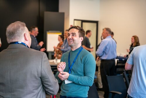 The image depicts a lively networking or professional event taking place in a modern conference room. The setting features a group of individuals engaged in conversation, suggesting a seminar, workshop, or business meeting. The room is well-lit with a neutral color palette, predominantly featuring whites and grays, contributing to a professional atmosphere.

In the foreground, a man wearing a teal sweater and a blue lanyard is smiling and actively engaging in conversation with another individual whose back is turned to the camera. This interaction suggests a friendly and open atmosphere. Other attendees are scattered throughout the room, some standing in small groups, indicating multiple conversations happening simultaneously.

Chairs and tables are visible, arranged to facilitate interaction, and a flip chart is positioned against the wall, hinting at presentations or collaborative activities. The attire of the participants is business casual, reinforcing the professional nature of the event.

The overall mood is positive and energetic, with attendees appearing engaged and interested. The image captures the essence of a collaborative and dynamic professional gathering, emphasizing networking and communication.