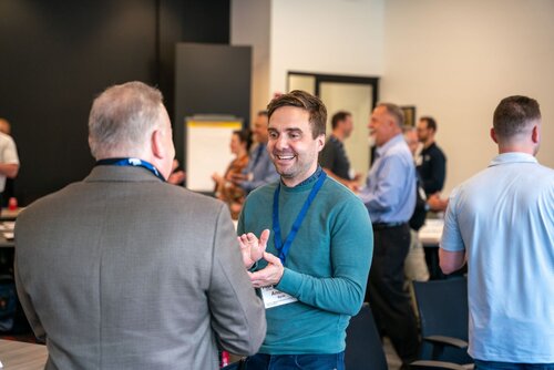 The image depicts a lively networking or professional event taking place in a modern conference room. The setting features a group of individuals engaged in conversation, suggesting a workshop, seminar, or business meeting atmosphere. 

In the foreground, two men are interacting warmly. One, facing the camera, is wearing a teal sweater and a blue lanyard with a name badge, smiling and gesturing with his hands, indicating an engaging and friendly conversation. The other man, with his back to the camera, is dressed in a brown suit jacket and also wears a lanyard, suggesting this is a formal or semi-formal event.

The background shows several other attendees, both men and women, involved in similar interactions, creating a bustling and energetic environment. A flip chart is visible, hinting at presentations or discussions taking place. The room is well-lit with a combination of natural and artificial lighting, contributing to an open and inviting atmosphere.

The predominant colors are neutral tones with pops of blue from the lanyards, which may signify event organizers or participants. The overall mood is professional yet relaxed, with an emphasis on networking and knowledge exchange. The image conveys a sense of collaboration and community within a professional setting.