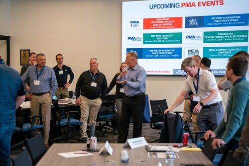 The image depicts a professional meeting or seminar taking place in a conference room. A group of individuals, mostly men, are gathered around tables, engaging in discussion. The setting is modern, with a clean and organized layout. The attendees are dressed in business casual attire, wearing name tags, indicating a formal event or networking session.

Prominently displayed on a large screen at the front of the room is a slide titled "UPCOMING PMA EVENTS." This slide lists several events with dates and locations, such as "Management Development Academy," "Deep Draw Technology Seminar," and "Metal Stamping Technology Conference." These events are associated with organizations like PMA (Precision Metalforming Association) and MDA (Management Development Academy), suggesting the meeting is related to the manufacturing or metalworking industry.

The atmosphere is professional and collaborative, with participants actively engaged in conversation, suggesting a focus on learning, development, or strategic planning. The room is well-lit, contributing to a focused and energetic environment. The presence of laptops, notepads, and a soda can on the tables indicates a working session where attendees are likely taking notes or accessing digital resources.

Overall, the image conveys a sense of industry-specific professional development, networking, and knowledge sharing, typical of conferences or seminars in the business sector.