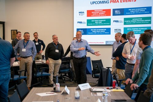 The image depicts a professional meeting or seminar setting with a group of individuals engaged in discussion. The room is equipped with tables and chairs arranged for a collaborative environment. On the tables, there are name tags, notebooks, and beverages, suggesting an organized event.

In the background, a large screen displays a slide titled "UPCOMING PMA EVENTS," listing several events with dates and locations. These include:

- Management Development Academy, Cleveland, OH
- Deep Draw Technology Seminar, Cleveland, OH
- One Voice In-District Public Policy Update
- Metal Stamping Processes Seminar, Cleveland, OH
- Metal Stamping Technology and Conference, Nashville, TN

The individuals, mostly men, are dressed in business casual attire, indicating a professional yet relaxed atmosphere. One man in the center appears to be speaking or presenting, suggesting he might be leading the discussion or providing information to the group.

The room is well-lit with natural and artificial lighting, creating a bright and focused environment. The overall atmosphere is one of collaboration and professional engagement, typical of industry conferences or training sessions.

The setting and content suggest a focus on manufacturing or metalworking industries, as indicated by the event titles related to metal stamping and technology seminars. The image conveys a sense of networking and knowledge sharing among industry professionals.