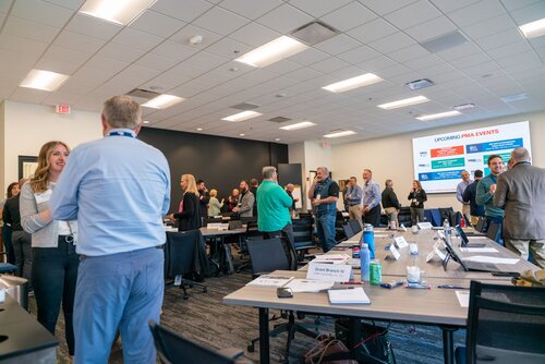 The image depicts a professional networking or conference event taking place in a modern, well-lit meeting room. The setting includes several tables arranged in a U-shape, with nameplates, notepads, and water bottles on them, suggesting a formal gathering. Attendees, both men and women, are casually dressed in business attire, engaging in conversations and networking activities. 

The room features a ceiling with recessed lighting, creating a bright and inviting atmosphere. A large screen at the front displays a slide titled "UPCOMING PMA EVENTS," indicating that this event is related to the PMA organization, possibly a professional association. The slide lists various events, though the details are not clearly visible.

The predominant colors in the room are neutral tones, with white walls and dark carpeting, complemented by the attendees' varied clothing colors. The atmosphere is professional yet relaxed, typical of a business or industry conference where networking and information exchange are key activities.

Overall, the image captures a dynamic and interactive environment, emphasizing professional engagement and collaboration.