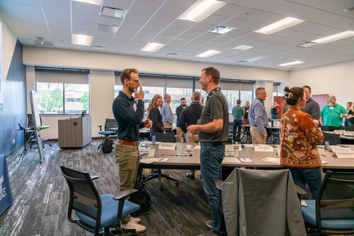 The image depicts a professional setting, likely a conference or workshop, held in a modern meeting room. The room is well-lit with overhead fluorescent lighting and large windows that allow natural light to enter, creating a bright and open atmosphere. The predominant colors are neutral tones, with shades of gray and blue on the walls and furniture, contributing to a professional ambiance.

Several individuals are engaged in conversation, suggesting a networking or collaborative event. They are casually dressed, indicating a relaxed yet professional environment. The tables are arranged in a U-shape, with name tags and water bottles placed on them, typical of a seminar or training session. Some people are standing, holding coffee cups, which adds to the informal and interactive nature of the gathering.

The room features modern office chairs and tables, and there is a visible projector screen, hinting at presentations or discussions taking place. The presence of a banner in the background, although not entirely legible, suggests branding or sponsorship, which is common in corporate events.

Overall, the image conveys a sense of professional engagement and collaboration, with an emphasis on interaction and communication among participants. The setting and activities are indicative of a business or educational context, focusing on networking and knowledge sharing.
