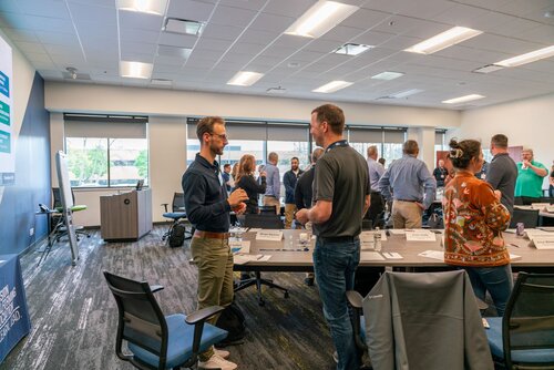 The image depicts a professional meeting or workshop taking place in a modern conference room. The setting features a large room with a high ceiling and ample natural light streaming in through large windows on one side. The room is furnished with long tables arranged in a U-shape, surrounded by ergonomic office chairs. 

Several individuals are engaged in conversation, suggesting a networking or collaborative session. The attendees are dressed in business casual attire, and they appear to be interacting in a relaxed yet focused manner. Nameplates are visible on the tables, indicating an organized event where participants may be identified by name.

On the left side of the image, a banner or poster is partially visible, featuring a logo and text that might relate to the organization hosting the event. The predominant colors in the room are neutral tones, with blue and gray being prominent in the furniture and decor, contributing to a professional and calm atmosphere.

The lighting is bright and evenly distributed, enhancing the clarity of the scene and emphasizing the professional environment. The overall atmosphere conveys a sense of collaboration, learning, and professional engagement, typical of corporate workshops or training sessions.