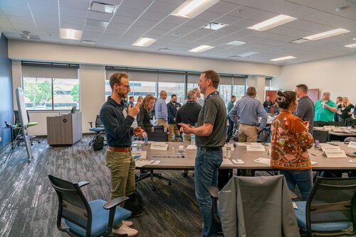 The image depicts a professional setting, likely a workshop or seminar, taking place in a modern conference room. The room is well-lit with natural light streaming in through large windows, complemented by overhead fluorescent lighting. The predominant colors in the room are neutral tones, with gray carpeting and a mix of black and gray furniture.

In the foreground, two men are engaged in conversation. One is wearing a dark shirt and khaki pants, while the other is in a gray polo shirt and jeans. Both appear to be animated and engaged, suggesting a lively discussion or networking session. Around them, several other individuals are standing and interacting in small groups, indicating a break or interactive session within the event.

The tables are arranged in a U-shape, with name tags and bottled water placed on them, suggesting an organized and formal event. A flip chart is visible to the left, indicating the presence of presentations or collaborative activities. The attendees are dressed in business casual attire, which aligns with the professional yet relaxed atmosphere of the event.

The overall atmosphere is one of collaboration and engagement, typical of a corporate training or team-building event. The presence of technology, such as laptops and projectors, suggests a modern and interactive environment. The setting and interactions imply a focus on learning, networking, and professional development.