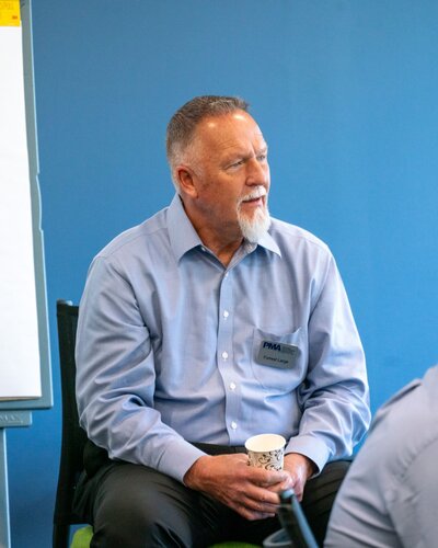 The image depicts a man seated in a casual, professional setting. He is wearing a light blue dress shirt and dark pants, and he has a white beard. He holds a paper cup with a decorative pattern in his left hand. The man appears to be engaged in conversation or listening attentively, as suggested by his focused expression and body language.

He has a name tag on his shirt with the text "PMA" and "Ernest L." The background is a solid blue wall, which adds a calm and neutral tone to the setting. To the left, part of a whiteboard is visible, indicating a meeting or workshop environment. The lighting is bright and even, suggesting an indoor setting with ample natural or artificial light.

The atmosphere is professional yet relaxed, typical of a corporate meeting or seminar. The presence of the name tag and whiteboard suggests a formal gathering, possibly for training or discussion purposes. The image conveys a sense of engagement and attentiveness, characteristic of collaborative or educational events.