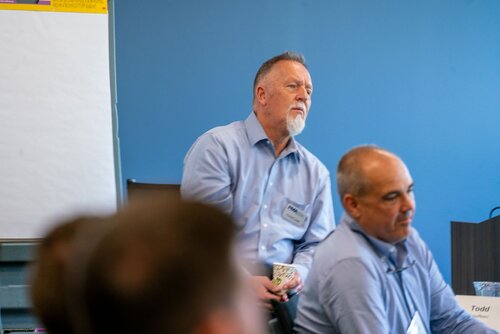 The image depicts a professional setting, likely a meeting or workshop, with a focus on two individuals. The man in the foreground, wearing a light blue shirt, is seated and appears to be listening attentively. He has a name tag that reads "Todd." Another man, also in a light blue shirt, is slightly behind him, standing and leaning against a table or desk. His name tag reads "Paul." Both individuals have a serious, contemplative demeanor, suggesting engagement in a discussion or presentation.

The background features a blue wall and a large flip chart or whiteboard, indicating a corporate or educational environment. The lighting is bright and even, typical of an indoor office space. The overall atmosphere is professional and focused, suggesting a business meeting or training session.

The image conveys a sense of attentiveness and participation, common in professional gatherings where information exchange and collaboration are key. The attire and setting suggest a formal or semi-formal business context.