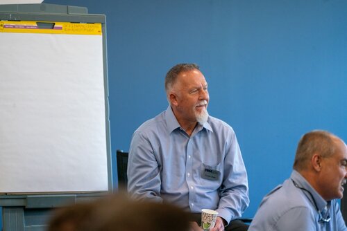 The image depicts a professional setting, likely a workshop or seminar. In the foreground, a man with a white beard and short hair is seated, wearing a light blue button-up shirt. He appears engaged and is holding a paper cup, suggesting a casual, interactive atmosphere. The man has a name tag, indicating a formal event where participants are identifiable.

To his right, another individual is partially visible, also wearing a similar shirt, suggesting a uniform or dress code for the event. The background features a large, blank flip chart on a stand, hinting at a presentation or brainstorming session. The flip chart has a yellow header, possibly for labeling or categorizing information.

The setting is well-lit, with a blue wall in the background contributing to a calm and focused environment. The overall atmosphere is professional yet relaxed, indicative of a collaborative or educational event. The image captures a moment of attentive listening or contemplation, typical of a learning or discussion-based scenario.