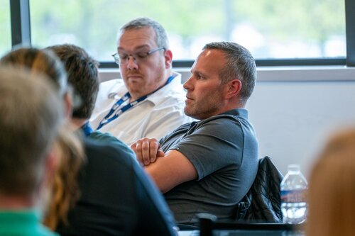 The image depicts a group of individuals seated indoors, likely participating in a meeting or conference. The setting appears to be a modern, well-lit room with large windows allowing natural light to enter, creating a bright and open atmosphere. 

In the foreground, two men are prominently visible. The man on the left is wearing glasses and a white shirt with a lanyard, suggesting a professional or corporate environment. The man on the right is dressed in a casual dark polo shirt and appears to be attentively listening, with his arms crossed. A plastic water bottle is placed on the table in front of him, indicating a casual or informal meeting setup.

The background shows blurred figures, emphasizing the focus on the two main individuals. The overall color palette is neutral, with a mix of natural light and indoor lighting contributing to a calm and focused atmosphere.

The setting and attire suggest a professional context, possibly a workshop, seminar, or team meeting. The image conveys a sense of attentiveness and engagement among the participants.