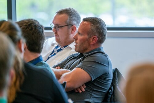 The image depicts a group of individuals seated in what appears to be a conference or meeting room. The focus is on two men in the foreground. The man on the right is wearing a dark polo shirt and has his arms crossed, suggesting a contemplative or attentive posture. The man next to him is wearing glasses and a white shirt, looking slightly downward, possibly engaged in listening or taking notes.

The setting is indoors, with a large window in the background that allows natural light to illuminate the room, creating a bright and open atmosphere. The window reveals a blurred view of greenery outside, indicating a daytime setting.

The room is filled with other attendees, as suggested by the presence of additional people visible from behind, though they are not the main focus. The attire of the individuals suggests a professional or semi-formal event, possibly a business meeting, seminar, or workshop.

The overall mood is one of focus and engagement, typical of a professional gathering where participants are likely discussing or learning about specific topics. The environment is conducive to concentration and interaction, with a neutral color palette that emphasizes the professional setting.