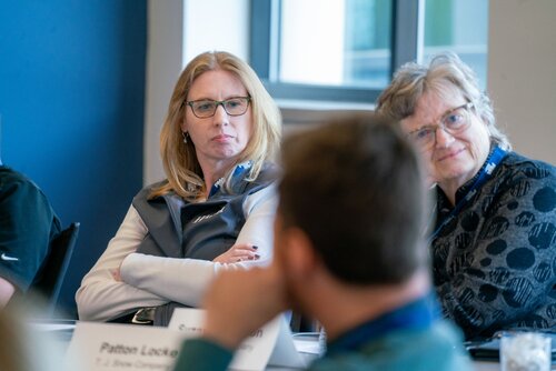 The image depicts a professional meeting or conference setting, with three individuals prominently featured. The setting appears to be a conference room with large windows allowing natural light to illuminate the space. The walls are painted in a muted blue, contributing to a calm and focused atmosphere.

In the foreground, a woman with long blonde hair and glasses is seated, wearing a gray vest over a white shirt. Her arms are crossed, and she appears to be attentively listening to someone off-camera. Next to her, another woman with short gray hair and glasses is also engaged, wearing a patterned dark top. Both women exhibit expressions of concentration and interest, suggesting a serious and thoughtful discussion.

A man is partially visible in the foreground, facing away from the camera, indicating he might be speaking or presenting to the group. He is wearing a blue shirt, and his posture suggests engagement with the conversation.

On the table, there are nameplates, one of which reads "Pattern Lock" followed by some text that is not fully visible. This detail suggests the meeting might involve discussions on technology or security topics.

The overall atmosphere is professional and focused, indicative of a formal business or academic meeting. The image captures a moment of active listening and exchange of ideas, common in collaborative environments.