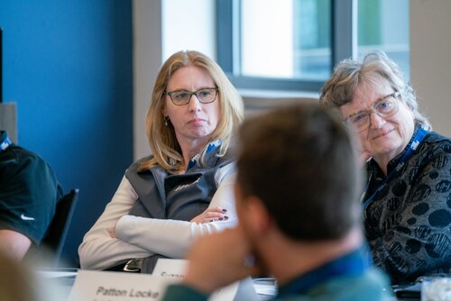 The image depicts a group of individuals engaged in what appears to be a meeting or conference setting. The scene is indoors, with a focus on two women seated at a table. The woman on the left has blonde hair, wears glasses, and is dressed in a gray vest over a white shirt. She appears attentive, with her arms crossed, suggesting engagement or contemplation. The woman on the right has gray hair, also wears glasses, and is dressed in a patterned dark top. She is smiling, which adds a warm and friendly atmosphere to the scene.

In the foreground, there is a man with his back to the camera, slightly blurred, indicating a depth of field that emphasizes the women. Nameplates are visible on the table, suggesting a formal setting, possibly a workshop or seminar. The environment is professional, with blue and white walls and a large window providing natural light, contributing to a bright and open atmosphere.

The image conveys a sense of collaboration and discussion, typical of professional gatherings. The overall mood is one of attentiveness and collegiality, with an emphasis on communication and interaction among participants.