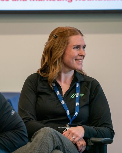 The image features a woman seated, wearing a black long-sleeve top with a logo on the chest that appears to read "TE3." She has light brown hair styled with a small clip. Around her neck is a blue lanyard with white text, though the specific text is not clearly visible. The woman is smiling and looking to her left, suggesting she might be engaged in a conversation or listening to someone speak.

The setting appears to be indoors, possibly a conference or meeting room, indicated by the formal attire and the presence of a lanyard, which is often used for identification at events. The background is neutral, with a portion of a screen or board visible at the top, displaying some text in red and black, though the text is not fully legible.

The lighting is bright and even, typical of indoor settings, contributing to a professional and positive atmosphere. The woman's expression and body language convey a sense of engagement and attentiveness, suggesting a professional or educational context.