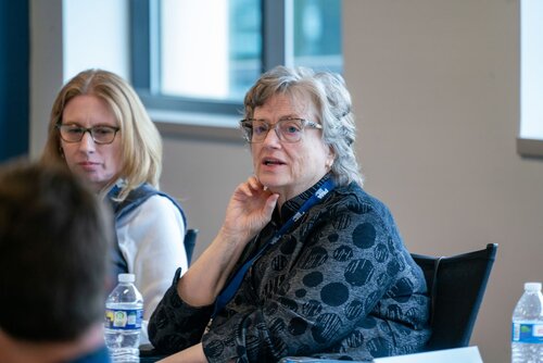The image depicts a meeting or discussion setting, likely in a conference room. Two individuals are prominently featured, both seated and engaged in conversation. The woman in the foreground, with gray hair and glasses, is wearing a dark, patterned blouse and a lanyard, suggesting a professional or academic event. She appears to be speaking or listening intently, with her hand resting near her chin, conveying an attentive and thoughtful demeanor.

The second woman, seated to her left, has blonde hair and is also wearing glasses. She is dressed in a light-colored top with a dark vest. Both women have water bottles in front of them, indicating a casual yet professional environment.

The room is well-lit with natural light coming through large windows in the background, creating a bright and open atmosphere. The setting suggests a formal meeting or seminar, possibly within an educational or corporate context.

The overall mood is professional and focused, with an emphasis on communication and engagement among participants. The presence of lanyards and the setting suggest a conference or workshop, emphasizing collaboration and discussion.