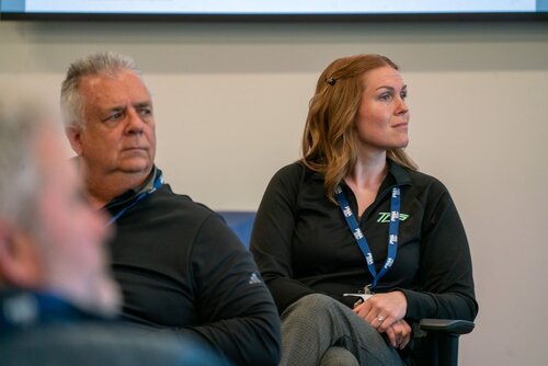The image depicts a professional setting, likely a conference or meeting room. Two individuals are prominently featured, seated and attentively listening to something off-camera. The man on the left has gray hair and is wearing a black zip-up jacket. The woman on the right has light brown hair and is also dressed in a black zip-up jacket, which features a logo with the letters "ZIE" visible. Both individuals are wearing lanyards with ID badges, suggesting a formal or organized event.

The background is neutral, with a light-colored wall and part of a projection screen visible at the top, indicating a presentation or discussion might be taking place. The lighting is bright and even, typical of indoor conference environments.

The atmosphere is focused and attentive, with the individuals appearing engaged in the proceedings. The setting suggests a business or educational context, possibly involving training, a seminar, or a corporate meeting. The presence of the logo and lanyards implies a professional or branded event.