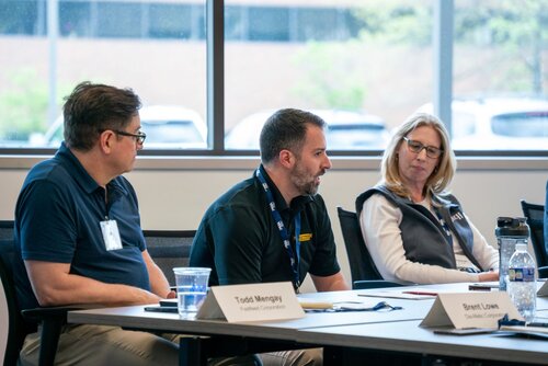 The image depicts a professional meeting or conference setting with three individuals seated at a table. The setting appears to be a modern conference room with large windows in the background, allowing natural light to illuminate the space. The individuals are engaged in a discussion, with one person speaking while the others listen attentively.

The man in the center, wearing a black polo shirt with a yellow logo, appears to be addressing the group. He has a focused expression, suggesting he is making a point or explaining something important. To his left, a man in a navy blue polo shirt with glasses listens intently, while to his right, a woman in a white shirt and black vest looks on thoughtfully.

On the table, there are nameplates identifying the individuals as Todd Mongay and Brent Lowe, suggesting a formal meeting context. There are also water bottles, a glass of water, and papers, indicating a typical setup for a business meeting or workshop.

The atmosphere is professional and collaborative, with an emphasis on communication and engagement. The setting and attire of the individuals suggest a corporate or educational environment. The overall tone is serious yet collegial, with a focus on discussion and exchange of ideas.