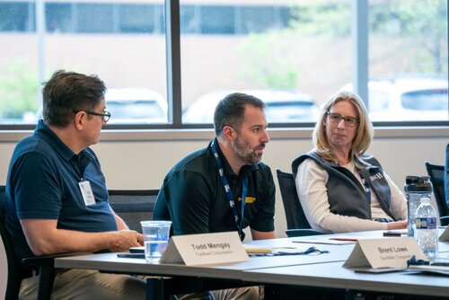 The image depicts a professional meeting or conference setting with three individuals seated at a table. The individuals appear engaged in a discussion. The setting is indoors, with natural light coming through large windows in the background, which also reveal a glimpse of parked cars outside.

1. **Individuals and Actions**:
   - The person in the center, wearing a black polo shirt with a yellow logo, appears to be speaking or responding to a question. He has a focused expression.
   - The individual on the left, wearing glasses and a dark blue shirt, is listening attentively, with a slight turn towards the speaker.
   - The person on the right, wearing a white shirt and a dark vest, is also listening, with a thoughtful expression.

2. **Text and Identifiable Objects**:
   - Name tags are visible on the table in front of the individuals. The tags read "Todd Mengel" and "Brent Lowe," along with their affiliations, "TrueNorth Companies" and "The HON Company," respectively.
   - A water bottle, a glass of water, and a notebook or document are on the table, suggesting a formal meeting environment.

3. **Colors and Lighting**:
   - The predominant colors are neutral tones, with black, white, and blue being prominent in the attire and setting.
   - The lighting is bright and natural, contributing to a professional and open atmosphere.

4. **Atmosphere and Context**:
   - The atmosphere is professional and collaborative, indicative of a business meeting or workshop.
   - The presence of name tags and company affiliations suggests a formal or semi-formal corporate event.

5. **Setting**:
   - The setting appears to be a conference room or office space, with modern furniture and a clean, organized layout.

Overall, the image conveys a sense of professional engagement and collaboration, with individuals actively participating in a business-related discussion.