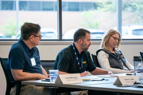 The image depicts a professional meeting setting with three individuals seated at a conference table. The setting appears to be a modern office or conference room, characterized by large windows in the background that allow natural light to illuminate the space. The individuals are engaged in discussion, with one person actively speaking while the others listen attentively.

The table is equipped with nameplates, suggesting a formal meeting or discussion. Two of the nameplates are visible, labeled "Todd Mengel" and "Brent Lowe," indicating their roles or affiliations, possibly with a company or organization. Papers, a pen, and a water bottle are also present on the table, typical of a business meeting environment.

The individuals are dressed in business casual attire, with polo shirts and vests, contributing to a professional yet relaxed atmosphere. The predominant colors in the image are neutral tones, with blues and grays being prominent, which adds to the formal and subdued ambiance.

The overall atmosphere conveys a sense of focus and collaboration, as the participants are engaged in what appears to be a structured discussion or presentation. The presence of nameplates and the formal setting suggest this could be a corporate meeting, workshop, or seminar, possibly involving strategic planning or project discussions.