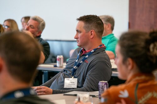 The image depicts a professional or educational setting, likely a conference or seminar. Individuals are seated at tables, attentively listening to a speaker or presentation. The focus is on a man wearing a gray jacket with a name badge that reads "Travis" and "Innovation" underneath. He is wearing a lanyard with a pattern of logos or symbols, suggesting affiliation with an organization or event.

In the background, other attendees are visible, with a mix of casual and business-casual attire, indicating a relaxed yet professional atmosphere. A few beverage cans are on the tables, hinting at a casual refreshment break or ongoing session. The lighting is bright and natural, typical of a conference room or meeting space.

The setting conveys a theme of learning and engagement, with participants focused on gaining knowledge or discussing innovative ideas. The atmosphere is one of concentration and attentiveness, typical of professional gatherings aimed at networking or skill development.