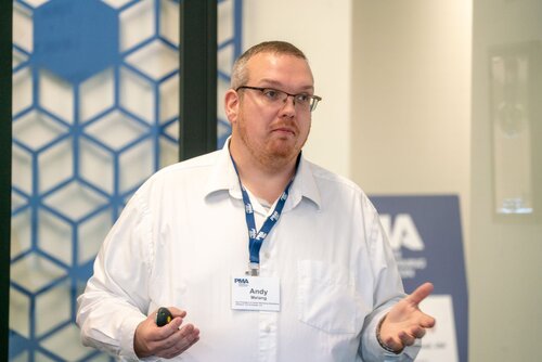 The image depicts a man standing indoors, possibly during a presentation or seminar. He is wearing a white shirt and glasses, with a lanyard around his neck that holds a name badge. The badge reads "Andy" and "McLoughlin," with "PMA" also visible, suggesting an association with a professional or industry event. He is holding a small device in one hand, likely a remote control for a presentation.

The background features a geometric blue and white pattern on a wall, adding a modern and professional atmosphere to the setting. There is also a blurred sign or board with the letters "PMA" visible, reinforcing the context of a formal event or conference.

The lighting is bright and even, typical of indoor conference settings, and the overall mood is professional and focused. The man appears to be engaged in explaining or discussing a topic, as indicated by his open hand gesture and attentive expression. The setting suggests a business or educational environment, possibly related to a specific industry or professional association.