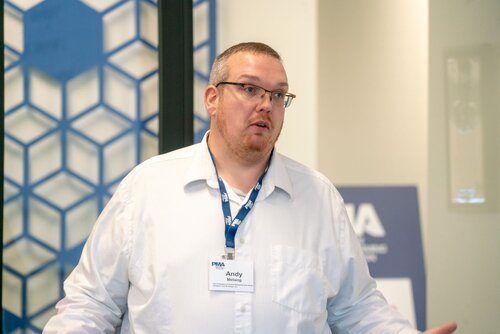 The image features a man wearing a white shirt and a blue lanyard with a name tag that reads "Andy." He appears to be speaking or presenting, with his hands slightly raised in a gesture. The background includes a geometric pattern with blue hexagons on a glass or transparent surface, contributing to a modern and professional setting. There is also a blurred sign in the background with the letters "PWA," suggesting a conference or professional event. The lighting is bright and even, indicating an indoor environment. The overall atmosphere is professional and focused, likely within a business or educational context.