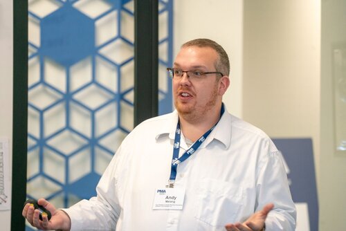 The image depicts a man, identified by a name tag as "Andy," who appears to be giving a presentation or speaking at an event. He is wearing a white shirt and a blue lanyard with a badge that reads "PMA." Andy is gesturing with his hands, suggesting he is actively engaging with an audience. The setting is likely a conference or seminar room, indicated by the professional attire and the presence of a geometric blue and white backdrop, which adds a modern and structured aesthetic to the scene. The lighting is bright and even, typical of indoor professional environments, contributing to a focused and formal atmosphere. The man's expression and body language convey a sense of confidence and engagement, suggesting he is knowledgeable about the topic he is discussing. The overall mood is professional and educational, suitable for a business or academic setting.