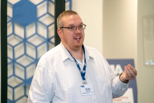 The image depicts a man engaged in a speaking or presentation activity, likely in a professional or conference setting. He is wearing a white shirt and a blue lanyard with a name badge that reads "Andy" and "Melrose." The badge also features the acronym "PMA," which might indicate the name of the organization or event.

The background includes a geometric pattern of hexagons in shades of blue, suggesting a modern or corporate environment. The lighting is bright and even, typical of indoor settings like conference rooms or offices.

The man appears to be gesturing with his right hand, possibly emphasizing a point or engaging with an audience. His expression suggests he is focused and involved in his presentation.

The atmosphere of the image is professional and educational, indicating a formal event such as a seminar, workshop, or business meeting. The presence of the name badge and lanyard reinforces the formal and organized nature of the event.

Overall, the image conveys a sense of professionalism and engagement, with a focus on communication and information sharing.
