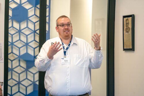 The image depicts a man standing indoors, engaged in a presentation or discussion. He is wearing a white shirt and glasses, with a lanyard around his neck that holds a name badge labeled "Andy." His hands are raised, suggesting he is emphasizing a point or explaining something to an unseen audience. The background features a blue and white geometric pattern resembling a honeycomb, which adds a modern and structured aesthetic to the setting. 

To the right, there is a framed portrait on the wall, contributing a formal or academic atmosphere to the environment. The lighting is bright and even, indicating a well-lit room, possibly an office or conference space. The overall mood of the image is professional and focused, suggesting a business or educational context. The presence of the name badge and lanyard implies a formal event, such as a conference, seminar, or workshop.