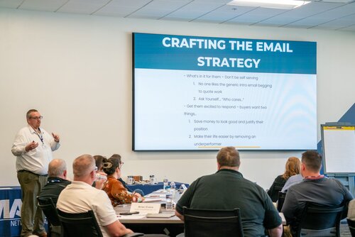 The image depicts a professional setting, likely a conference or workshop, focused on email marketing strategies. A speaker, dressed in a white shirt and dark pants, stands to the left, gesturing towards a large presentation screen. The screen displays the title "CRAFTING THE EMAIL STRATEGY" in bold, uppercase letters. Below the title, there are bullet points discussing tactics such as avoiding generic email content, making emails work for the recipient, and encouraging responses.

The audience consists of several individuals seated at tables, attentively listening and taking notes. They are dressed in business casual attire, suggesting a formal yet relaxed atmosphere. Nameplates and notebooks are visible on the tables, indicating an organized and structured event.

The room is well-lit with natural and artificial lighting, creating a bright and focused environment. The walls are plain, with a flip chart positioned next to the screen, hinting at interactive elements in the presentation.

The overall atmosphere is educational and professional, aimed at enhancing participants' skills in email marketing. The setting and content suggest a corporate training session or a seminar dedicated to improving business communication strategies.