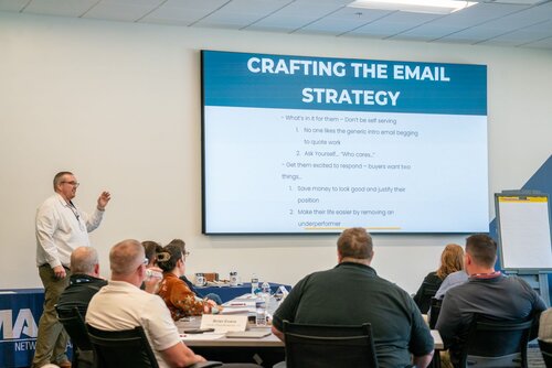 The image depicts a professional setting, likely a conference or workshop, focused on email marketing strategies. A man stands at the front of a room, gesturing towards a large presentation screen displaying the title "CRAFTING THE EMAIL STRATEGY." The slide includes bullet points discussing strategies such as avoiding generic emails, using humor, and appealing to buyers' interests.

Several individuals are seated at tables, facing the presenter, and appear to be engaged in the session. The attendees are diverse in terms of gender and attire, suggesting a casual business environment. On the tables are laptops, notebooks, and water bottles, indicating active participation and note-taking.

The room is well-lit with natural and artificial lighting, creating a bright and focused atmosphere. The walls are adorned with a banner that partially reads "MA..." suggesting a possible company or event name. An easel with a blank pad stands to the side, likely for additional notes or brainstorming.

The setting conveys a theme of professional development and learning, with an emphasis on practical skills in email marketing. The atmosphere is collaborative and educational, aimed at enhancing participants' understanding and effectiveness in crafting email strategies.