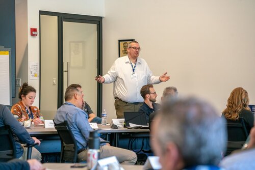 The image depicts a professional setting, likely a conference or workshop, with a group of individuals seated around tables. A man, standing at the front, appears to be giving a presentation or leading a discussion. He is dressed in business casual attire, wearing a white shirt and holding a remote or pointer, suggesting he is interacting with a presentation or slideshow.

The room is well-lit with natural and artificial light, creating a neutral and focused atmosphere. Attendees are seated, some taking notes or looking at laptops, indicating engagement with the content being presented. The presence of name tags and lanyards suggests a formal event, possibly involving networking or educational activities.

In the background, a glass door and a wall-mounted picture or poster are visible, contributing to the professional office or conference room setting. The attendees are diverse in appearance, reflecting a typical business environment.

The predominant colors are neutral tones, with a mix of white, black, and gray, complemented by the occasional splash of color from clothing and accessories. The overall mood is one of concentration and learning, typical of a corporate or educational event.