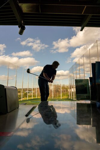 The image depicts a person engaged in a golf swing at an indoor driving range. The setting is a modern golf facility with a partially open roof, allowing a view of the sky filled with scattered clouds. The individual is silhouetted against the bright outdoor light, emphasizing their posture and the motion of the swing. 

In the foreground, a reflective surface captures the silhouette and adds depth to the composition. The scene is bathed in natural light, creating a contrast between the indoor shadows and the vibrant outdoor scenery. The background features a series of tall netting or barriers, typical of driving ranges, with a landscape of trees and a distant building visible beyond.

The atmosphere is dynamic and focused, capturing the concentration and precision associated with golfing. The setting suggests a recreational or practice environment, possibly at a facility like Topgolf, known for combining entertainment with sports. The overall mood is one of leisure and athleticism, set against a backdrop of clear skies and open space.