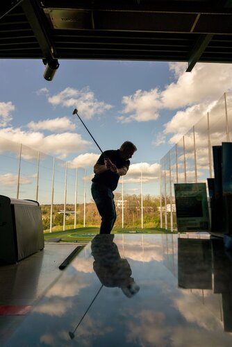 The image depicts a man engaged in a golf swing at an indoor driving range. He is silhouetted against a backdrop of a clear blue sky with scattered white clouds, visible through large, open windows or netted barriers that surround the range. The man is positioned centrally, mid-swing, with a golf club extended behind him, suggesting motion and focus.

In the foreground, a reflective surface, possibly a table or counter, mirrors part of the scene, adding depth and a dynamic visual element. The lighting is natural, with sunlight casting soft shadows and highlighting the man's silhouette, enhancing the contrast between the indoor setting and the bright outdoor environment.

The setting conveys a leisurely and focused atmosphere, typical of a recreational sports environment. The presence of modern equipment and the open-air design suggest a contemporary, possibly urban location. The image captures a moment of concentration and skill, typical of golf as a sport, and evokes a sense of calm and precision.