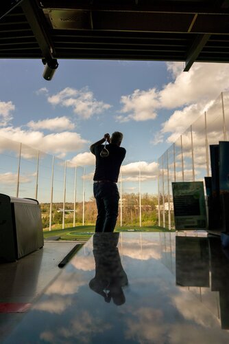 The image depicts a person engaged in a golfing activity at a driving range. The individual is captured mid-swing, facing away from the camera, with a golf club raised in the follow-through position. The setting is an open-air driving range, characterized by tall netting that surrounds the area, designed to catch golf balls. The sky is bright and partly cloudy, suggesting a clear day, which enhances the vibrant and energetic atmosphere of the scene.

In the foreground, there is a reflective surface, possibly a table, which mirrors the sky and part of the indoor structure. This reflection adds an artistic element to the composition. The lighting is natural, with sunlight illuminating the scene, casting soft shadows and highlighting the greenery in the background.

The predominant colors are the blues of the sky, the greens of the grass, and the neutral tones of the driving range structure. The overall mood is one of leisure and focus, typical of a recreational sports setting. The image conveys a sense of motion and concentration, capturing the essence of the golfing experience.