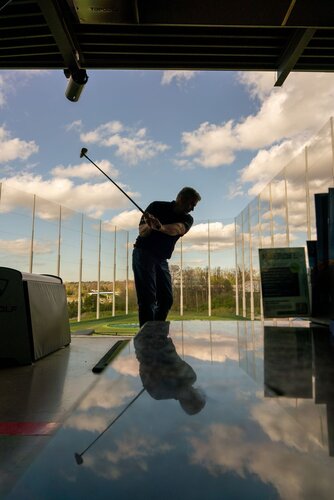 The image depicts a person engaged in a golf swing at an indoor driving range. The individual is silhouetted against a backdrop of a bright blue sky with scattered white clouds, creating a striking contrast. The golfer is positioned in the center, with their back slightly arched and arms extended, holding a golf club mid-swing. 

The setting is a modern golf facility, likely a Topgolf venue, indicated by the presence of a branded golf bag on the left. The area is open to the outside, with protective netting visible in the background, suggesting a controlled environment for practicing golf. The ground is reflective, possibly due to a polished surface, mirroring the golfer's silhouette and adding depth to the composition.

The lighting is natural, with sunlight illuminating the scene, enhancing the vibrant colors of the sky and the lush greenery visible in the distance. The overall atmosphere is dynamic and focused, capturing the concentration and motion of the sport.

The image conveys a sense of leisure and sport, typical of recreational golf settings, and emphasizes the blend of indoor comfort with outdoor elements. The combination of the golfer's action, the expansive sky, and the modern facility creates a contemporary and energetic mood.