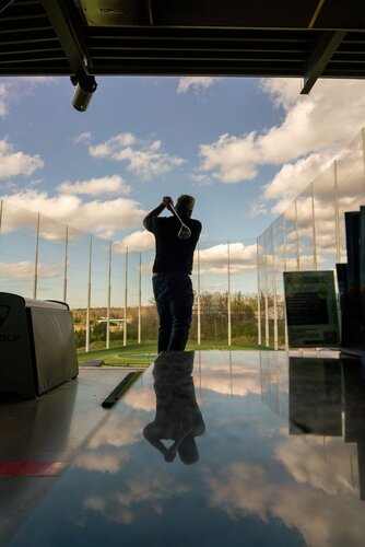 The image depicts a person playing golf at an indoor driving range. The individual is captured in mid-swing, silhouetted against a backdrop of a clear blue sky with scattered white clouds. The setting appears to be a modern golf facility, as indicated by the presence of a large netted enclosure surrounding the range, designed to catch golf balls.

In the foreground, there is a reflective surface, possibly a table, mirroring the sky and clouds above, adding a dynamic visual element to the composition. The lighting is natural, with sunlight casting soft shadows, suggesting a daytime setting.

The overall atmosphere is calm and focused, typical of a leisurely sports activity. The setting conveys a sense of relaxation and enjoyment, with the open sky contributing to a feeling of freedom and space.

This image might evoke themes of recreation and sportsmanship, commonly associated with golf. The presence of modern amenities suggests a contemporary setting, likely appealing to enthusiasts of the sport.