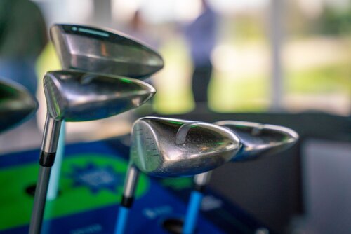 The image features a close-up of several golf clubs with metallic heads, prominently displayed in the foreground. The clubs are positioned upright, suggesting they are part of a set, possibly in a golf bag. The metallic sheen of the club heads reflects light, emphasizing their polished surfaces. In the background, there is a blurred view of individuals, possibly in conversation or observing, indicating a social or professional setting related to golf, such as a clubhouse or pro shop.

The setting appears to be indoors with large windows, allowing natural light to illuminate the scene. The predominant colors include the silver of the clubs and a mix of green and blue from what seems to be a surface or mat underneath them, adding a vibrant contrast to the metallic tones.

The atmosphere conveyed is one of leisure and sport, possibly highlighting themes of golfing culture or lifestyle. The presence of people in the background, though out of focus, suggests a communal or event-like environment, possibly a golf tournament or a demonstration event.

Overall, the image captures the essence of golf equipment in a setting that blends both the sport's technical aspects and its social dimensions.