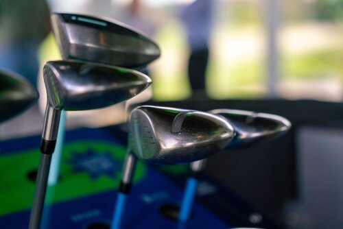 The image features a close-up of several golf clubs, specifically drivers, positioned upright in a golf bag. The clubs have shiny metallic heads with visible grooves, reflecting ambient light, which suggests a well-lit environment. The shafts of the clubs are blue, adding a splash of color against the metallic sheen of the club heads.

In the background, there is a blurred view of what appears to be a golf course or a driving range, indicated by the presence of greenery and a large window or open area allowing natural light to flood in. The lighting is bright and natural, contributing to a fresh and dynamic atmosphere.

The setting suggests a sporting environment, possibly a golf shop, a clubhouse, or a training facility. The presence of multiple clubs indicates preparation for a game or practice session. The overall theme conveys a sense of leisure and sportsmanship, typical of golfing activities.

The image captures the essence of golf as a sport, highlighting the equipment used and the serene, focused atmosphere associated with golfing environments.