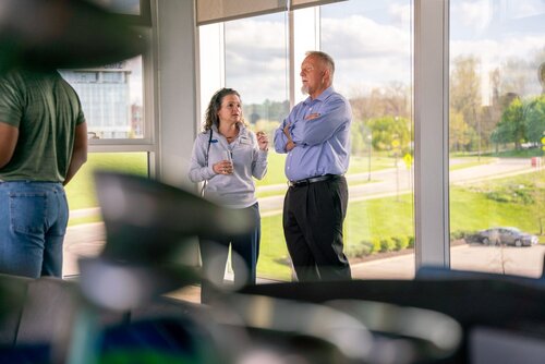 The image depicts a casual indoor setting with large windows offering a view of a green, sunny landscape outside. Two individuals are engaged in conversation near the windows. The woman on the left is wearing a light-colored jacket and holding a drink, while the man on the right is dressed in a blue shirt and dark pants, standing with his arms crossed. Their expressions suggest a friendly and engaged discussion.

The background shows a bright, clear day with lush greenery and a road visible through the windows, adding a sense of openness and tranquility to the scene. The lighting is natural, with sunlight streaming in, creating a warm and inviting atmosphere.

In the foreground, there are blurred objects, possibly furniture or decorative elements, which frame the scene and add depth to the image. The overall mood is relaxed and professional, indicating a setting that could be a casual business meeting or a networking event. The attire of the individuals suggests a business-casual environment.

The setting and attire imply a modern office or conference space, possibly within a corporate or educational institution. The image captures a moment of interpersonal connection, highlighting communication and engagement in a professional context.