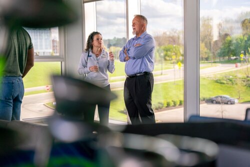 The image depicts a professional setting, likely within a modern office or conference room with large windows offering a view of a green, landscaped area outside. Two individuals are engaged in conversation. The woman on the left is holding a drink and appears to be speaking, while the man on the right, dressed in a blue shirt and dark pants, listens with his arms crossed. The atmosphere is casual yet professional, suggesting a business meeting or networking event.

The room is well-lit with natural light streaming in from the windows, creating a bright and open ambiance. The setting outside the windows includes a road and a grassy area, contributing to a serene backdrop. In the foreground, there are blurred objects, possibly furniture or decor, adding depth to the image.

The attire of the individuals suggests a business-casual environment. The image conveys a theme of communication and professional interaction, typical of a workplace or corporate event. The overall mood is relaxed yet focused, indicative of a productive and engaging discussion.