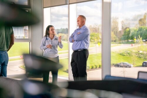 The image depicts two individuals engaged in conversation near a large window, offering a view of a green outdoor landscape. The setting appears to be a modern office or conference room, suggested by the large glass windows and the presence of blurred office equipment in the foreground. The man, dressed in a light blue shirt and dark trousers, stands with his arms crossed, listening attentively. The woman, wearing a light gray sweater and dark pants, gestures with her hands, indicating she is speaking or explaining something.

The lighting is bright and natural, streaming in from the window, which adds a warm and open atmosphere to the scene. The outdoor view includes a well-maintained lawn, trees, and a road, contributing to a serene and professional environment.

The overall mood is professional and collaborative, suggesting a business meeting or casual workplace discussion. The attire of the individuals and the setting indicate a formal or semi-formal context, possibly within a corporate or educational institution. The image conveys themes of communication, teamwork, and a modern work environment.