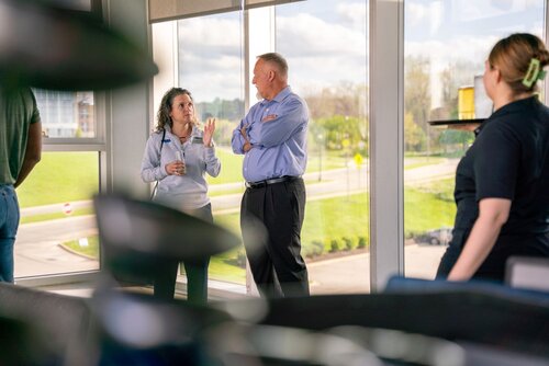 The image depicts a professional setting, likely an office or conference room with large windows offering a view of a green, outdoor landscape. Two individuals are engaged in conversation near the windows. The woman on the left, wearing a light gray sweater, appears to be explaining something, gesturing with her hands. The man on the right, dressed in a light blue shirt and dark pants, listens attentively with his arms crossed.

In the foreground, partially blurred, are two other individuals. One is a woman with her back to the camera, wearing a black shirt and with her hair tied back. The other person is obscured, with only parts of their clothing visible, suggesting they are seated or standing at a different angle.

The lighting is natural and bright, with sunlight streaming through the windows, creating a warm and open atmosphere. The setting suggests a collaborative or informal meeting, possibly discussing work-related topics or planning.

The overall mood is professional yet relaxed, indicative of a modern workplace environment where open communication is encouraged. The presence of greenery outside the windows adds a touch of tranquility and connection to nature, enhancing the positive ambiance of the scene.