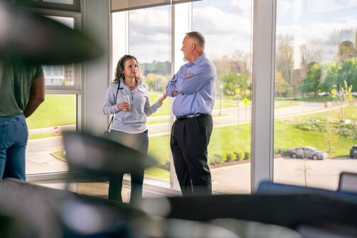 The image depicts a professional setting, likely within an office or conference space, characterized by large windows that offer a view of a suburban or park-like area outside. The lighting is bright and natural, suggesting it is daytime. Two individuals are engaged in conversation near the windows. 

The woman on the left is holding a drink, possibly a cup of coffee or tea, and is dressed casually in a light-colored sweater and dark pants. She appears to be actively listening or discussing something with the man on the right. The man is wearing a light blue dress shirt and dark trousers, exuding a business-casual appearance. His posture suggests he is explaining or responding to the woman.

In the background, the windows reveal a green landscape with trees and a road, indicating a peaceful and open environment outside. A few parked cars are visible, adding to the suburban or corporate campus feel.

The atmosphere is professional yet relaxed, suggesting a break or informal meeting. The presence of natural light and greenery outside contributes to a calm and open ambiance. The setting and attire imply a workplace or networking event, possibly during a break in a conference or meeting.