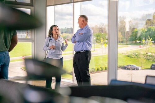 The image depicts a casual office setting with two individuals engaged in conversation near large windows. The woman on the left is holding a cup and gesturing with her other hand, suggesting an informal discussion. She is wearing a light-colored hoodie and dark pants. The man on the right, dressed in a light blue shirt and dark trousers, is listening attentively with his arms crossed.

The background reveals a bright, sunny day with a view of a green landscape, including trees and a road, visible through the expansive windows. The lighting is natural and bright, creating a warm and open atmosphere. The setting appears to be a modern office or conference room, characterized by its large windows and open view.

The emotional atmosphere is relaxed and conversational, indicative of a professional yet informal interaction. The overall theme suggests a collaborative or brainstorming session, typical in a business or educational environment. The image conveys a sense of openness and communication, emphasizing a positive and engaging work culture.