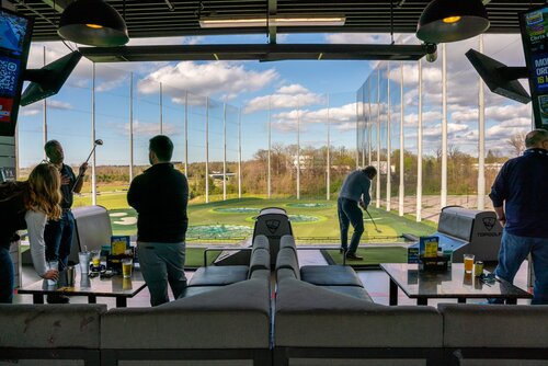 The image depicts an indoor golf entertainment venue, specifically a Topgolf location. The setting is a spacious, open-air driving range with individual bays for players. The foreground features a seating area with tables, where drinks are placed, suggesting a social and relaxed atmosphere. The venue's logo, "Topgolf," is visible on the furniture and digital screens, reinforcing the brand identity.

Several individuals are engaged in golfing activities. One person is swinging a golf club, while others watch or prepare to take their turn. The background shows a large, open field with targets marked on the ground, typical of a driving range designed for both practice and entertainment.

The scene is well-lit with natural daylight, highlighting a clear blue sky with scattered clouds, contributing to a vibrant and energetic ambiance. The overall mood is casual and recreational, appealing to both golf enthusiasts and those seeking a fun social outing.

The setting suggests a modern, leisure-focused environment, popular for group gatherings and events, blending sports with social interaction.