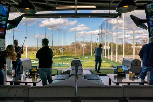 The image depicts an indoor golf entertainment venue, specifically a Topgolf location. The setting features a large, open driving range with multiple targets on a well-maintained green field. The area is enclosed by tall netting, and the sky is clear with scattered clouds, suggesting a pleasant day.

In the foreground, several individuals are engaged in playing golf. They are positioned in individual bays, each equipped with golf clubs and seating areas. Drinks are placed on tables, indicating a casual and social atmosphere. The players are in various stages of swinging their clubs, capturing the dynamic nature of the activity.

The interior is modern, with a sleek design. Overhead lighting provides a warm ambiance, complementing the natural light streaming in from outside. The seating is comfortable, with plush couches and tables, emphasizing a relaxed environment.

The presence of the Topgolf logo on the furniture and digital screens displaying information or advertisements adds to the branded experience. The overall atmosphere is lively and recreational, catering to both golf enthusiasts and those seeking a fun, social outing.